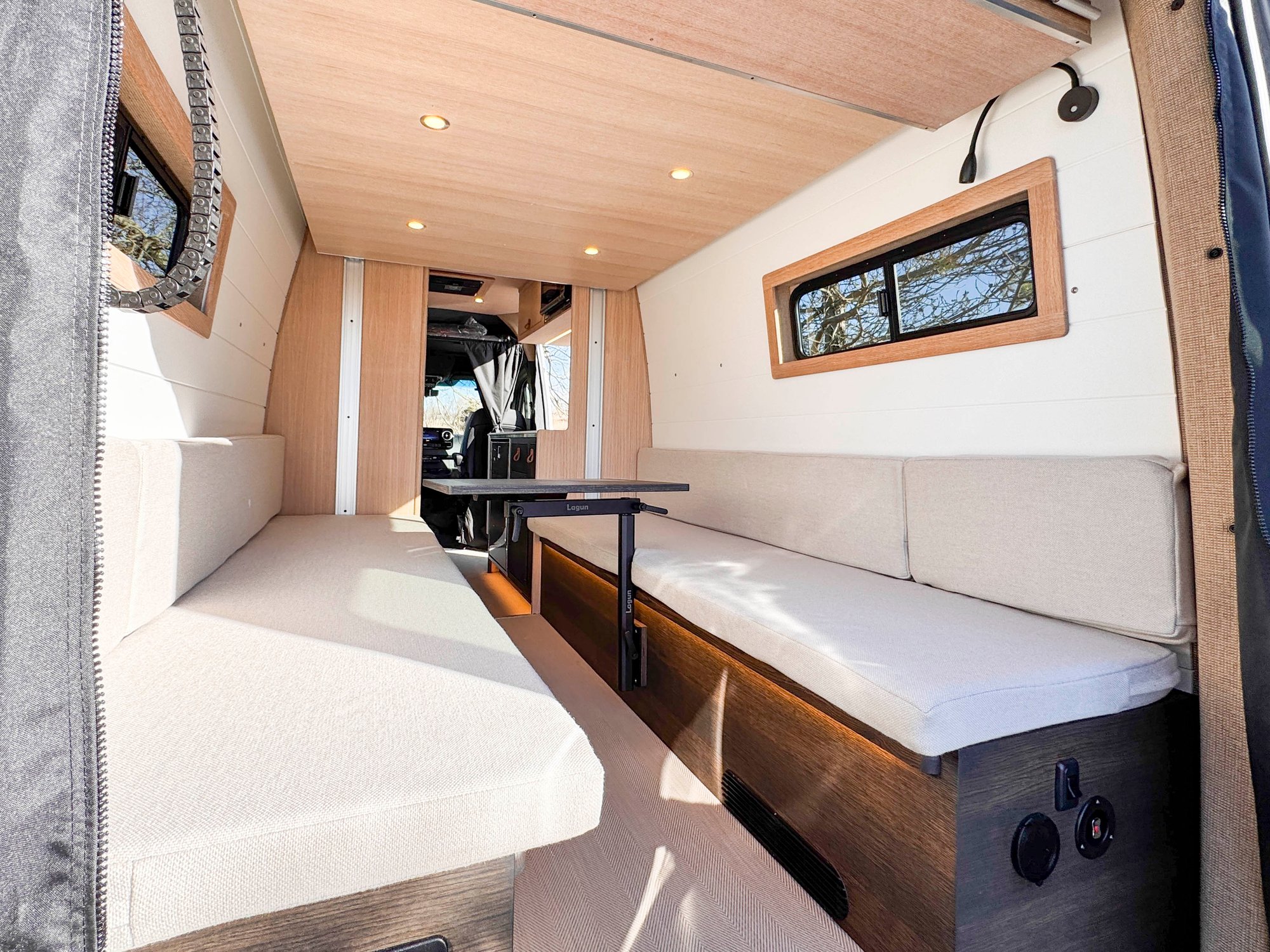 The Loft interior in day mode — long open cabin with dual oatmeal lounge benches and a Lagun table, looking forward toward the cab on the Sprinter 170