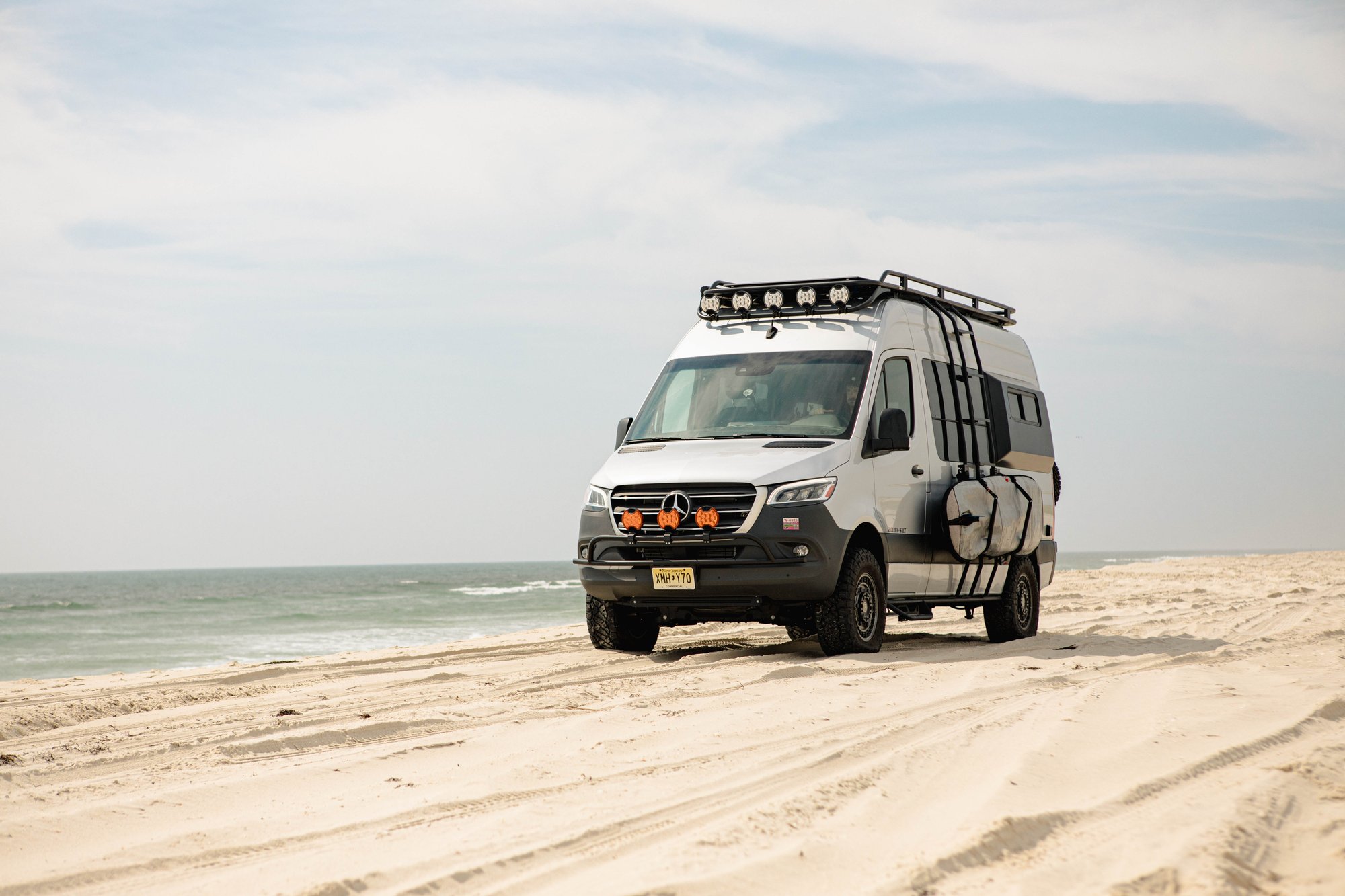 The Stoke — silver Sprinter 144 loaded with a surfboard on the side rack, parked on the beach