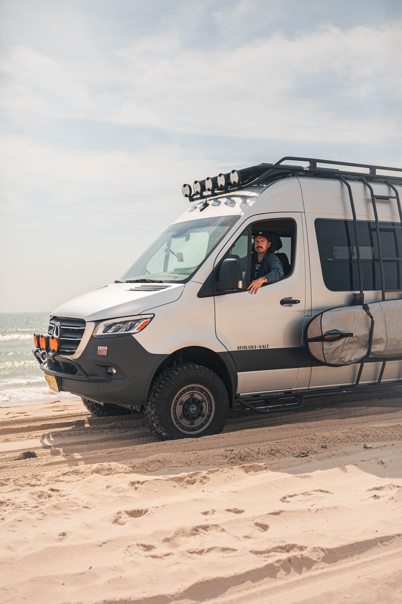 The Stoke — owner looking out the Sprinter 144 window with a surfboard, parked on the beach