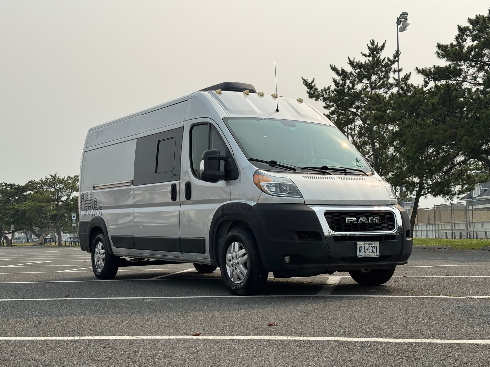 Silver 2019 Ram ProMaster 159 high-roof camper van, front three-quarter view in a beachside parking lot