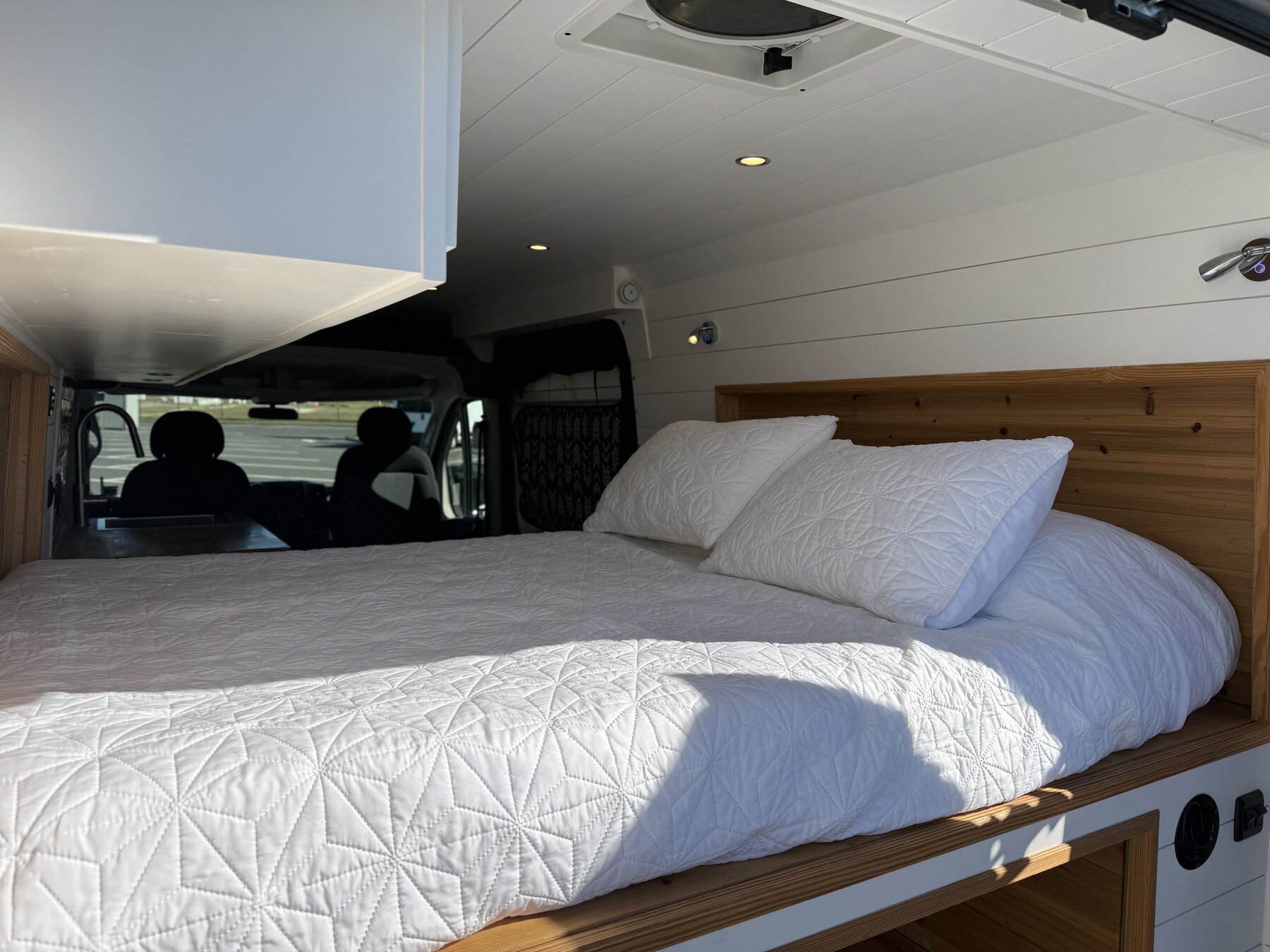 Bed seen from the front of the van, with overhead cabinets and a roof vent fan above