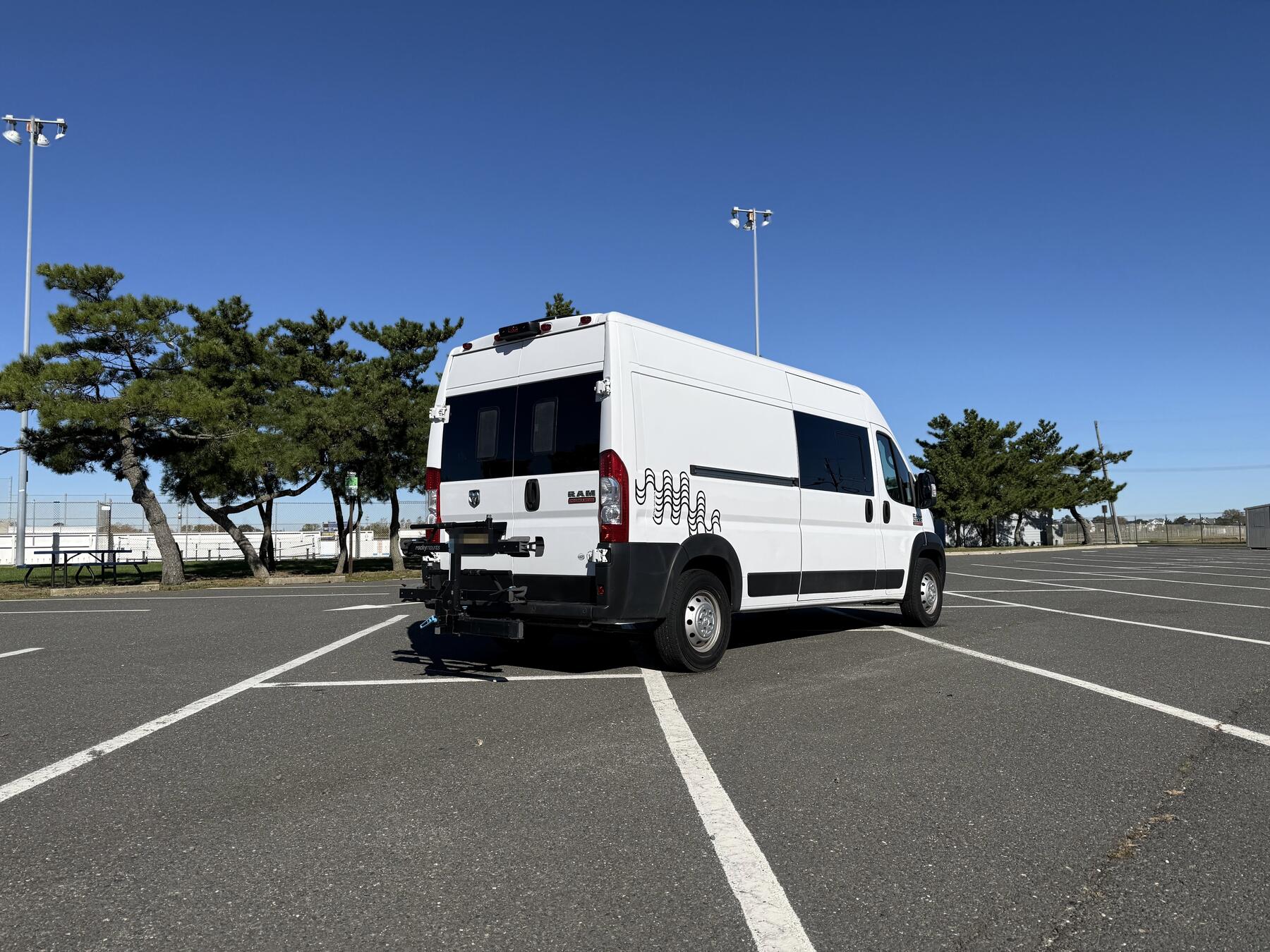 White Ram ProMaster high-roof camper van, rear three-quarter view in a parking lot with pine trees and blue sky