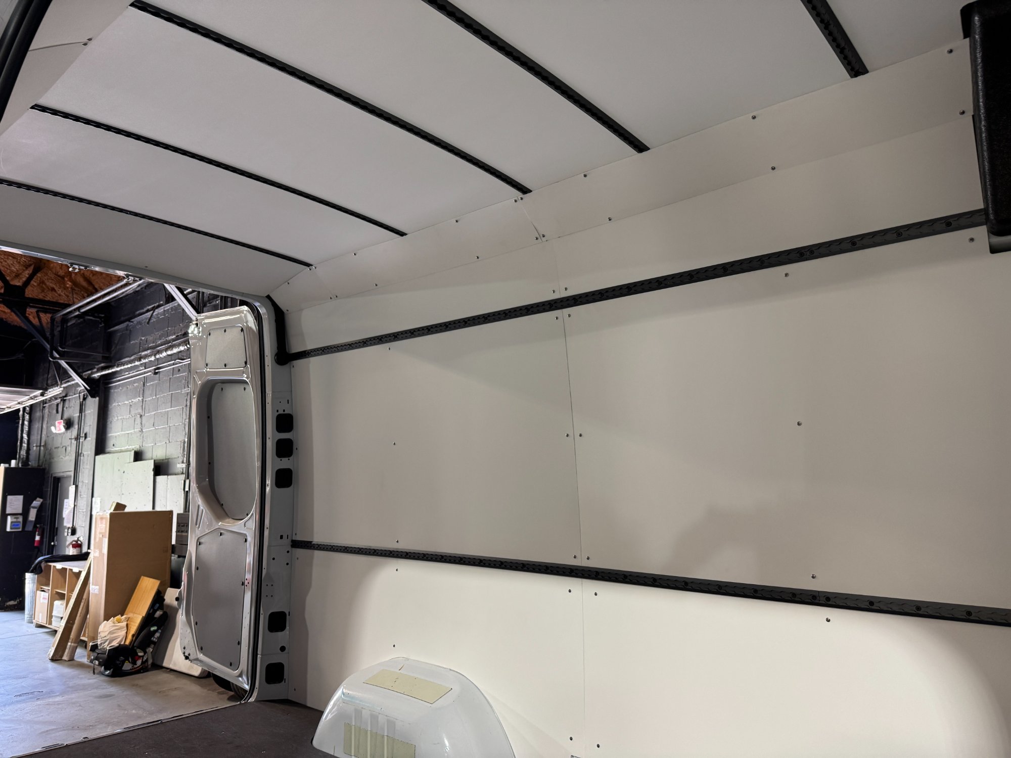 Sprinter conversion mid-Build phase — walls and ceiling fully paneled with mounting tracks installed, side door open in the Sequoia + Salt shop before cabinetry goes in