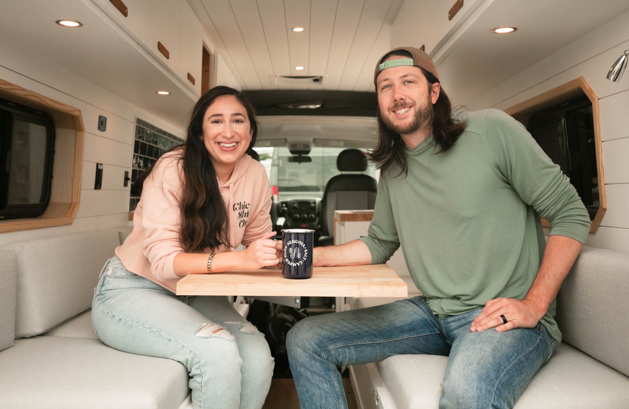 A Sequoia + Salt owner couple at their van's dinette with a Sequoia + Salt branded mug — the customers we build for